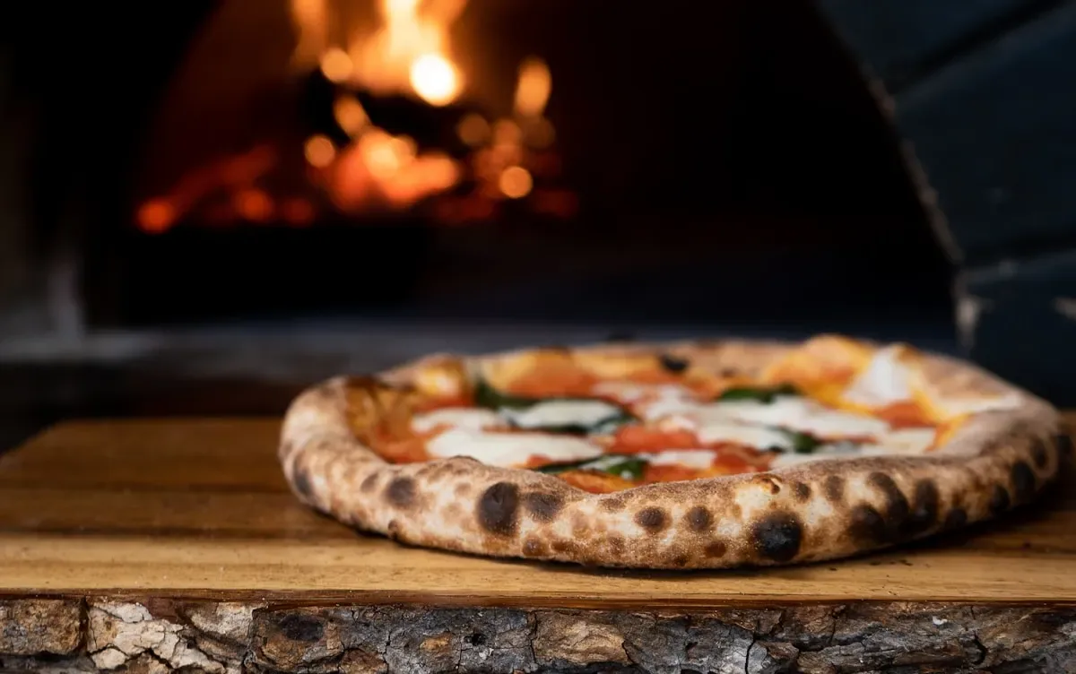 La bocca del forno a legna acceso, con una pizza sul piano di legno